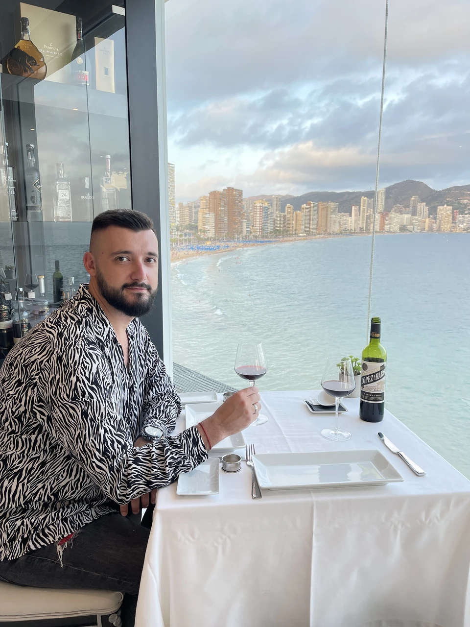 Артём Gameleon в тёплом свитере стоит на чёрном песчаном пляже Tjørnuvík у подножия зелёных крутых скал