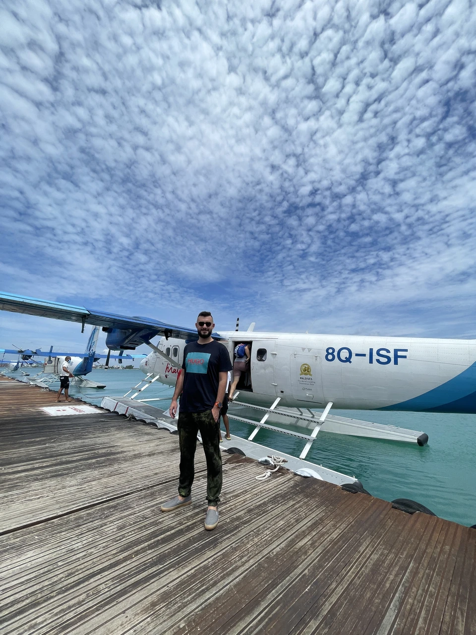 Артём Gameleon в куртке сидит на краю смотровой площадки и любуется величественным норвежским фьордом с зелёными склонами, снежными вершинами и драматичным облачным небом.