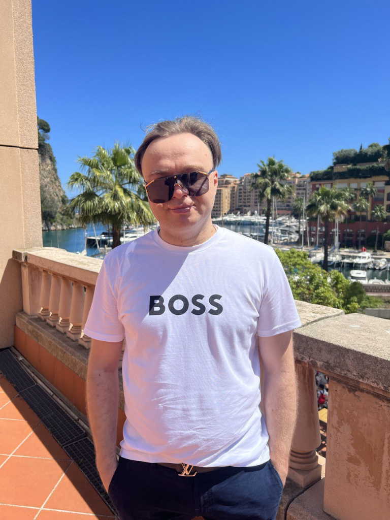 Артём Gameleon в белой футболке BOSS стоит на балконе с видом на яхтенный порт Монако, пальмы и небоскрёбы на фоне