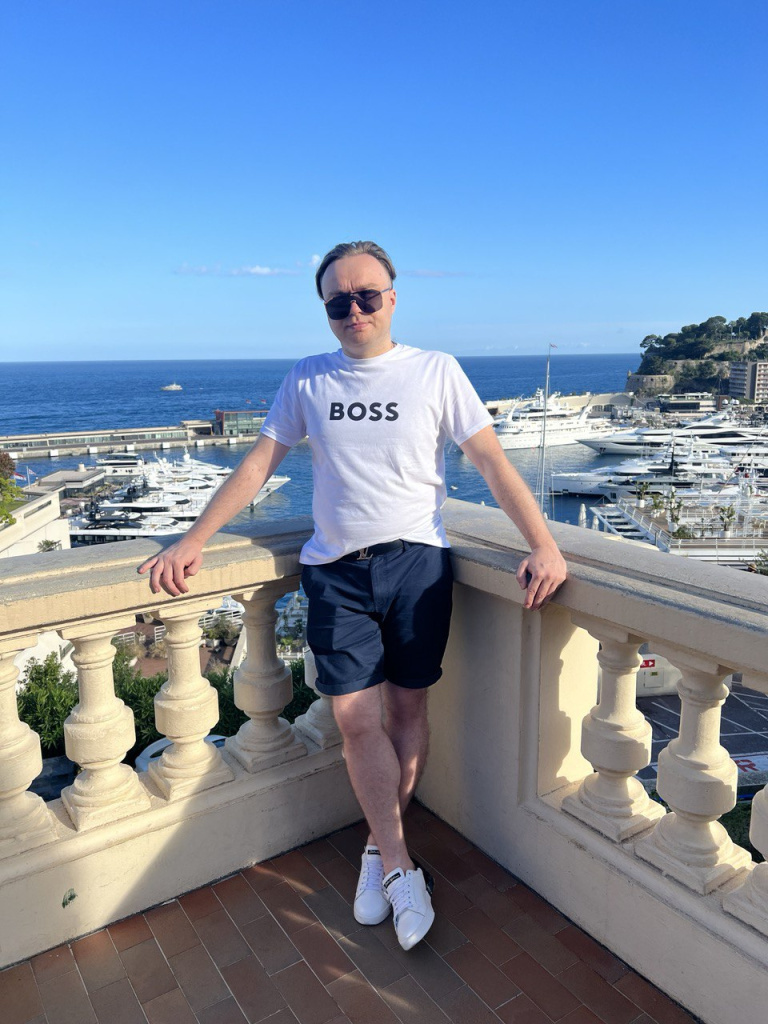 Артём Gameleon в белой футболке BOSS опирается на балюстраду с видом на причаленные яхты в порту Монако под ясным небом